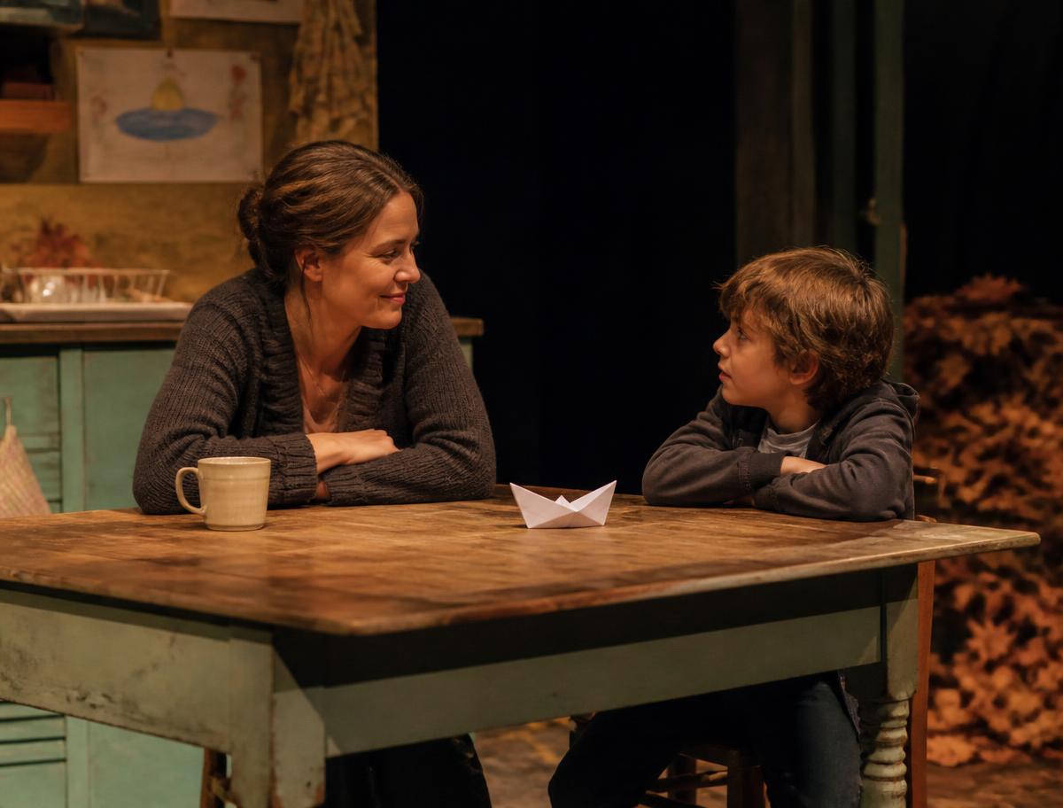 Two performers at a kitchen table with a folded paper boat between them, warm low stage light
